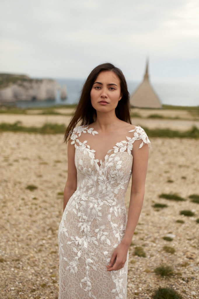Eine Frau in einem weißen Spitzenhochzeitskleid steht draußen am Strand mit Klippen und einem spitzen Gebäude im Hintergrund und zeigt nachhaltige Brautkleider und Pre-loved Traumkleid Inspiration für Brautmode in Stuttgart.