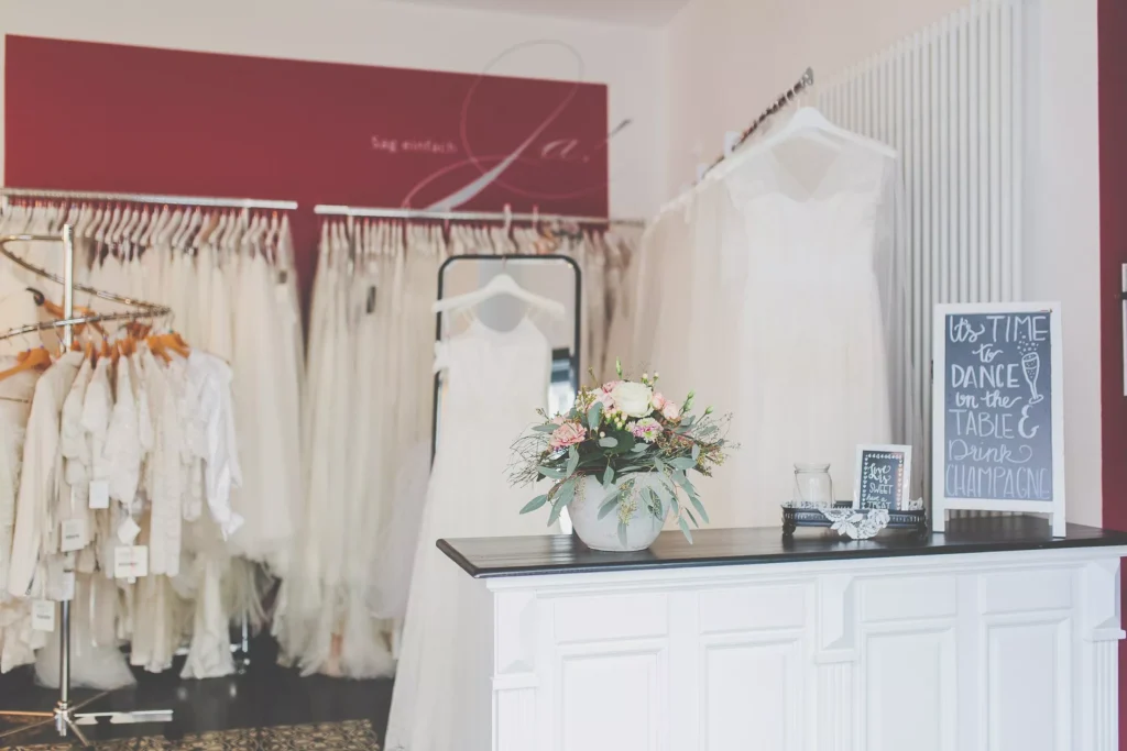 Ein Stuttgarter Brautmodengeschäft mit nachhaltigen Brautkleidern: weiße Brautkleider in Regalen, ein blumengeschmückter Tresen und ein charmantes Kreideschild. Entdecken Sie Ihr gebrauchtes Traumkleid mit Stil.
