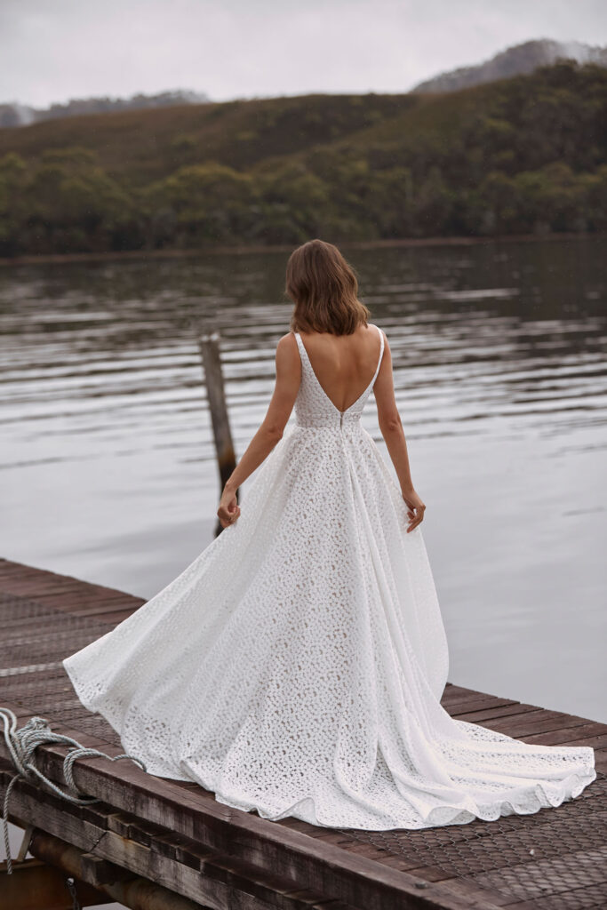 Eine Frau in einem weißen, spitzenbesetzten, rückenfreien Kleid - vielleicht ein Pre-loved Traumkleid - steht auf einem hölzernen Steg an einem ruhigen See und schaut in die Ferne.