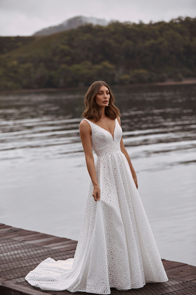 Eine Frau in einem ärmellosen weißen Hochzeitskleid steht auf einem Steg an einem ruhigen See mit bewaldeten Hügeln und trägt ein wunderschönes Pre-loved Traumkleid von Second Hand Brautkleider-perfekt für alle, die einzigartige Brautmode in Stuttgart suchen.