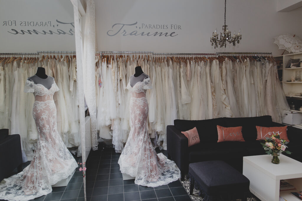 Ein Brautladen mit einem Spitzenhochzeitskleid auf einer Schaufensterpuppe, einer verspiegelten Wand und Kleiderständern hinter einem schwarzen Sofa - mit Second Hand Brautkleidern für Bräute, die ihr gebrauchtes Traumkleid in einem eleganten Rahmen suchen.