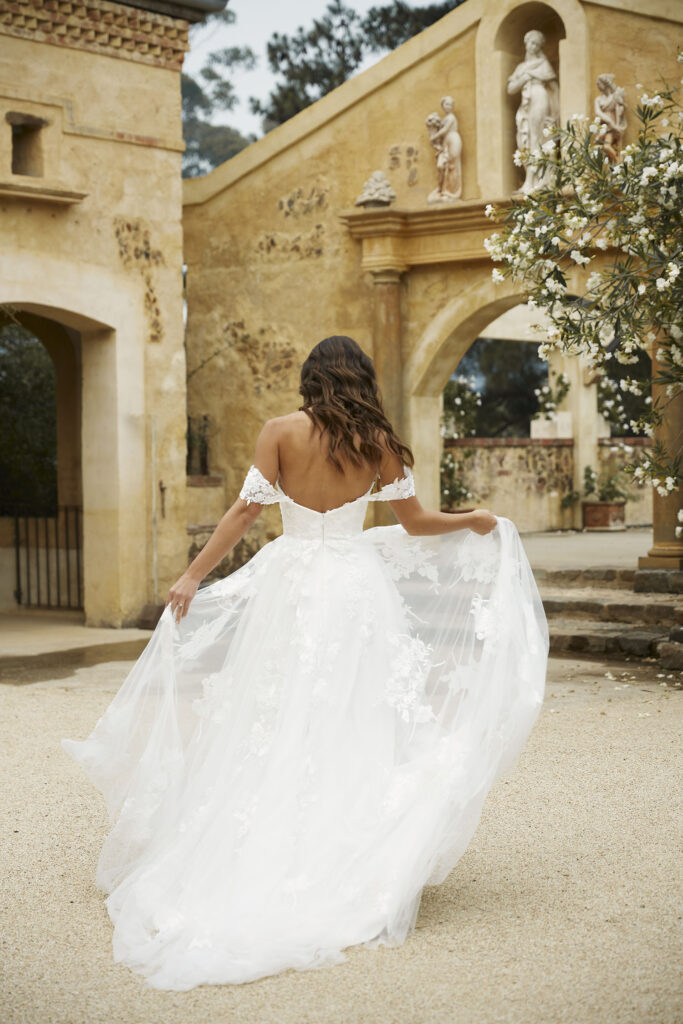 Eine Frau in einem schulterfreien weißen Hochzeitskleid steht im Freien und hält ihr Kleid in einem Garten mit Steinbögen - die perfekte Inspiration für nachhaltige Brautkleider und Brautmode in Stuttgart.