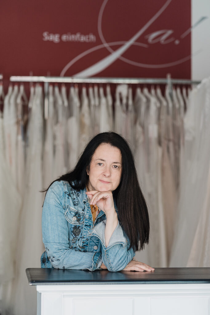 Doris mit langen dunklen Haaren in Jeansjacke lehnt an einem Tresen, mit nachhaltigen Brautkleidern und eleganten Hochzeitskleidern im Hintergrund.