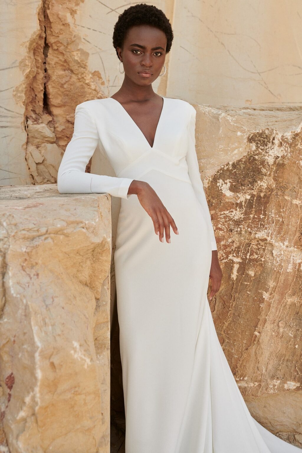 Eine Frau in einem langärmeligen weißen Kleid steht vor großen Steinplatten mit einem neutralen, strukturierten Hintergrund - die perfekte Inspiration, um nachhaltige Brautkleider oder Ihr eigenes Pre-loved Traumkleid zu entdecken.