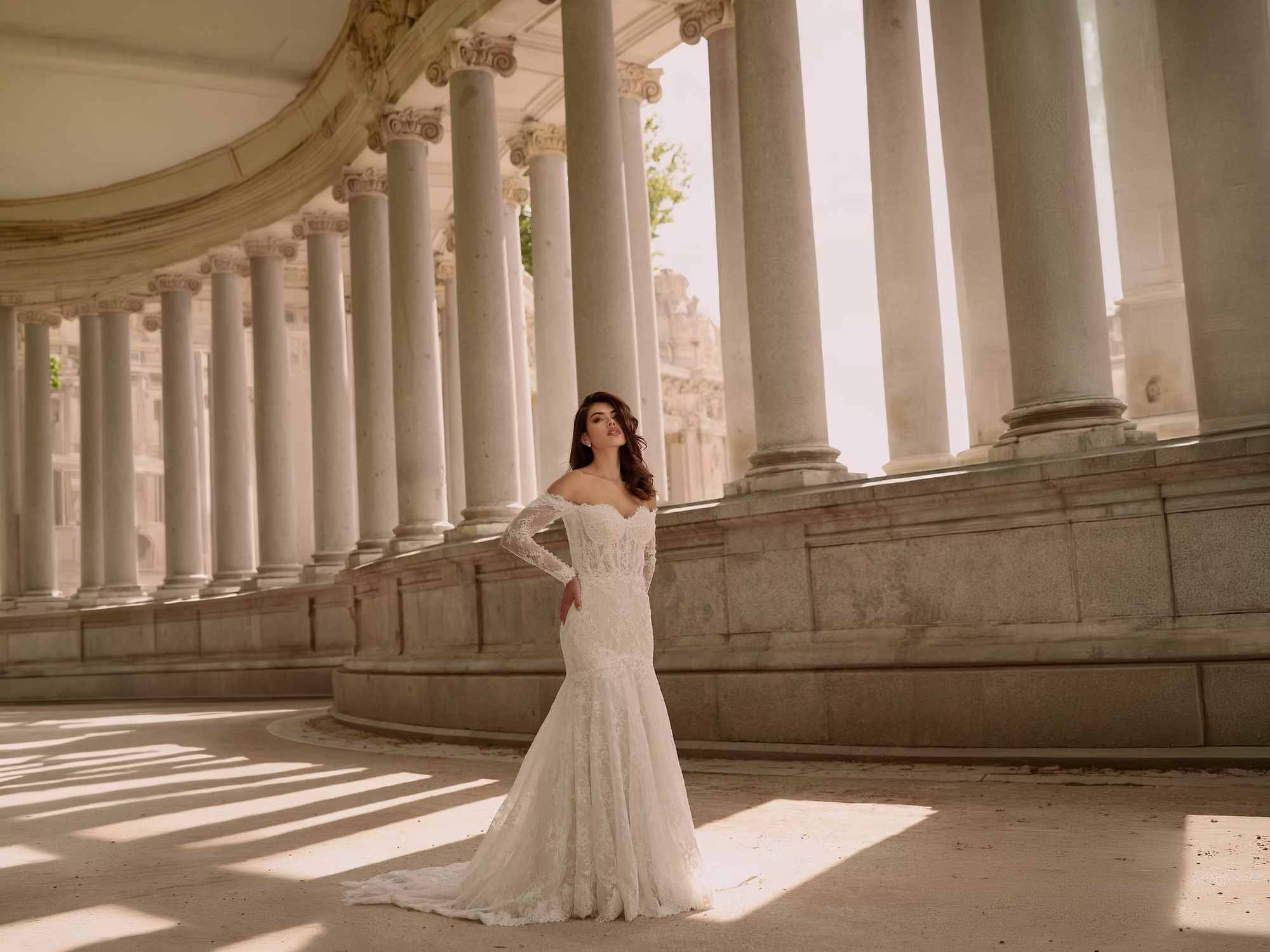Eine Frau in einem Second Hand Brautkleid aus weißer Spitze steht an Steinsäulen im Sonnenlicht und nimmt eine selbstbewusste Pose ein.