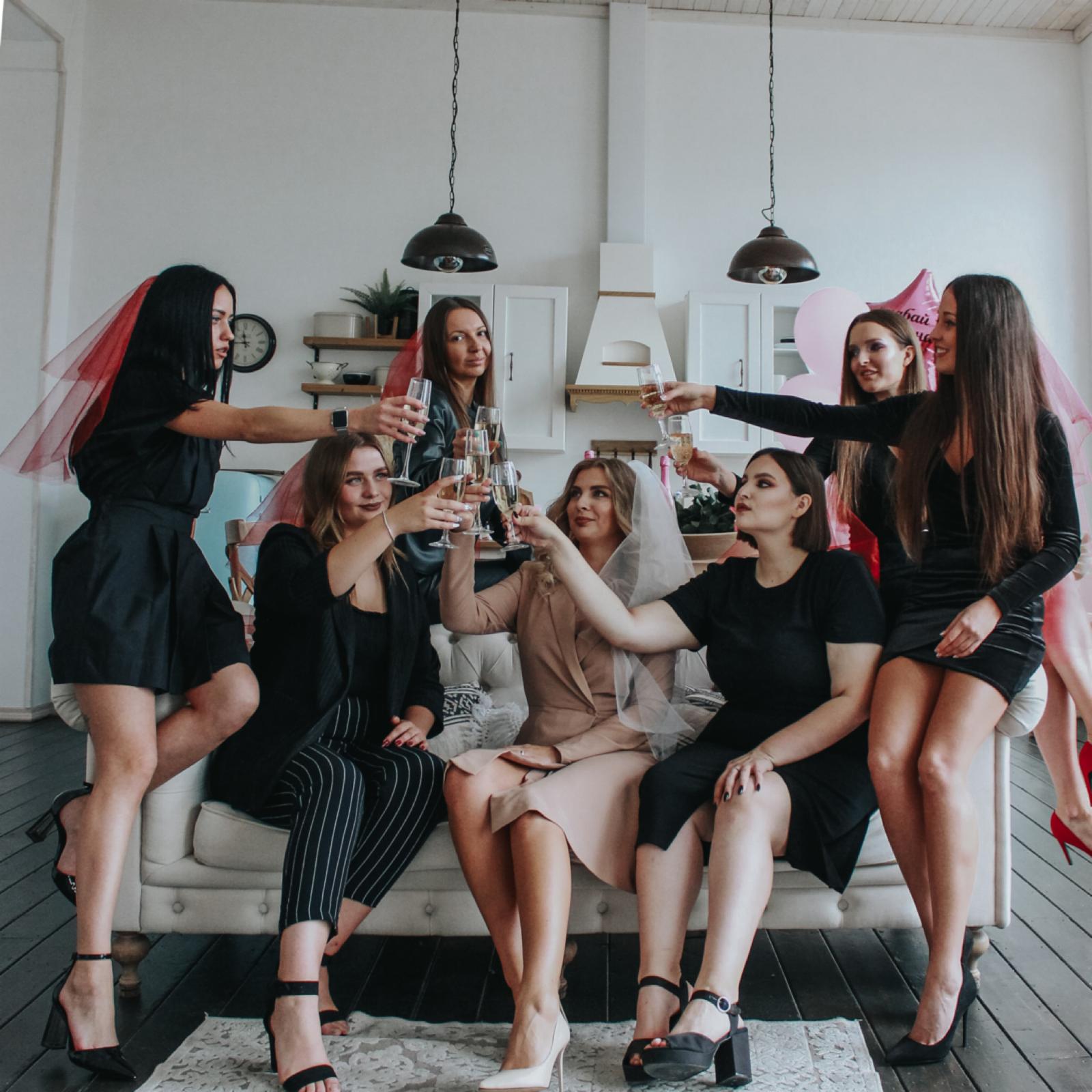 Sieben Frauen mit Schleiern und Partyoutfits, einige mit nachhaltigen Brautkleidern, stoßen an, während sie um eine Couch herum sitzen und stehen.