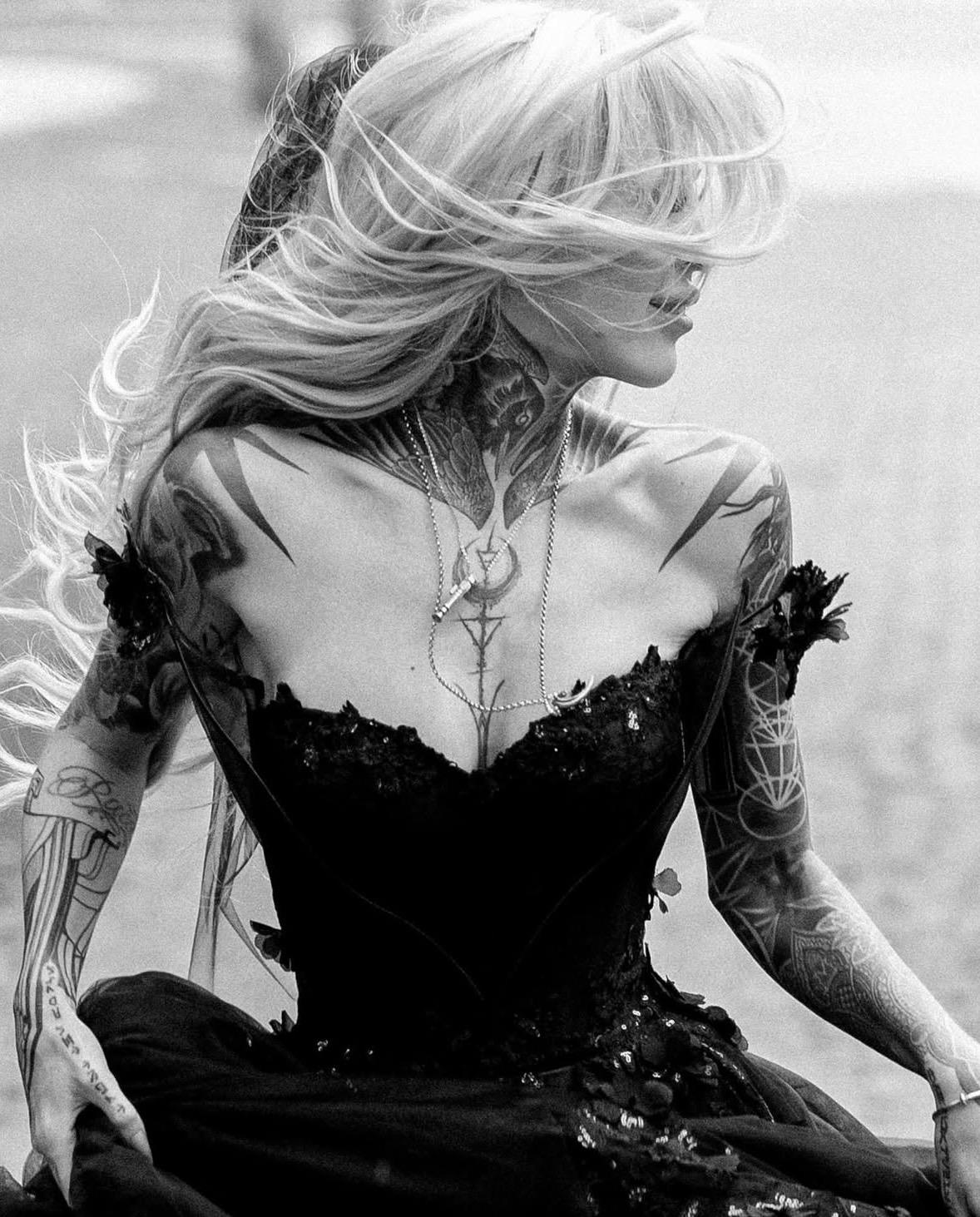 Eine tätowierte Frau in einem schwarzen Brautkleid mit vom Wind zerzausten Haaren, die auf einem Schwarz-Weiß-Foto zur Seite blickt.